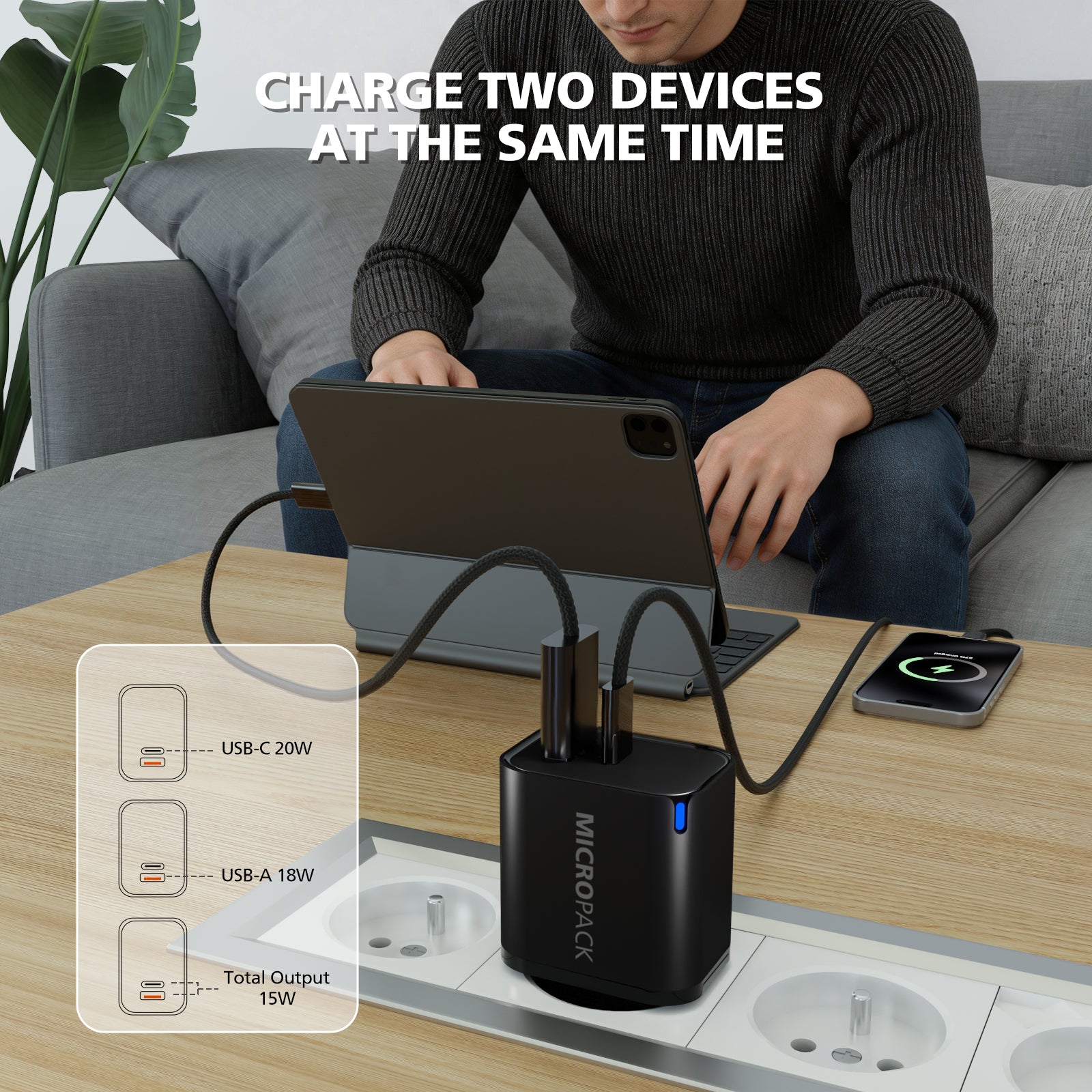 A black MICROPACK dual-device charger sits on a wooden table in a living room setting, with a man sitting on a gray sofa in the background charging a tablet and a smartphone. White text at the top reads 'CHARGE TWO DEVICES AT THE SAME TIME', and a diagram on the bottom left shows USB-C 20W, USB-A 18W, and total output 15W.