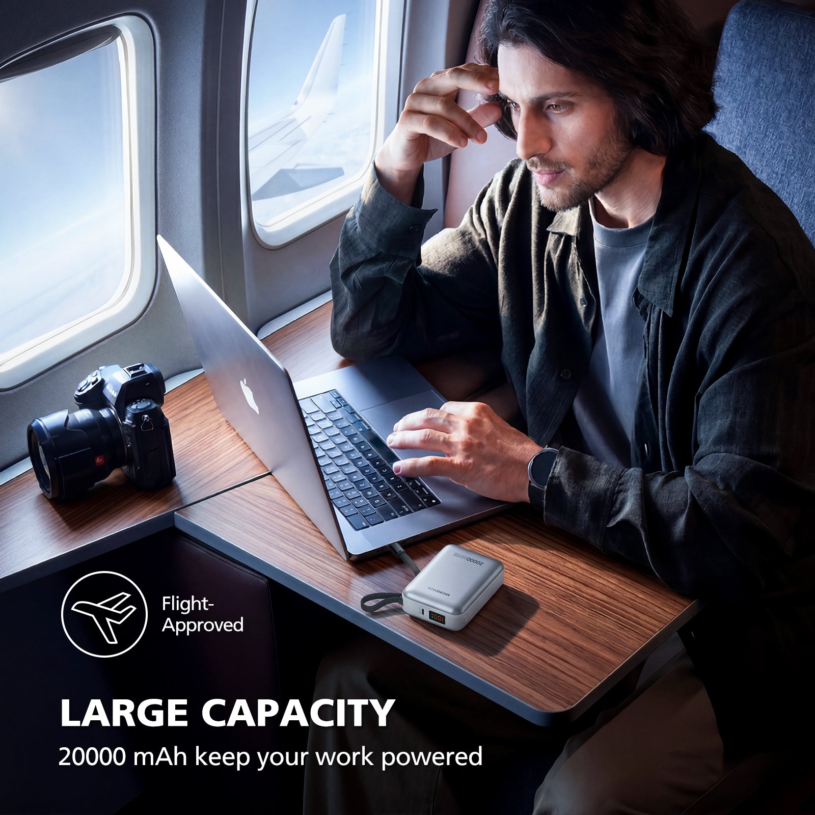 Man using a laptop on an airplane with a power bank and camera on a tray table.